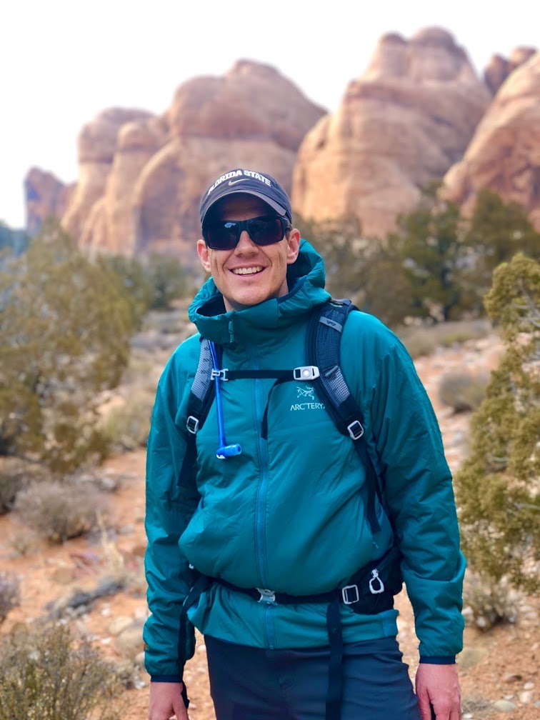 Smiling hiker in teal jacket and Florida State cap standing in a desert landscape with sandstone formations, symbolizing outdoor adventure, personal reflection, and connection to nature