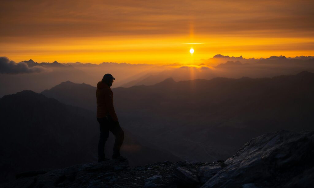 Silhouetted figure standing on a rocky mountain peak at sunrise, surrounded by glowing clouds and layered ridges—symbolizing reflection, achievement, and the journey of intentional fatherhood.