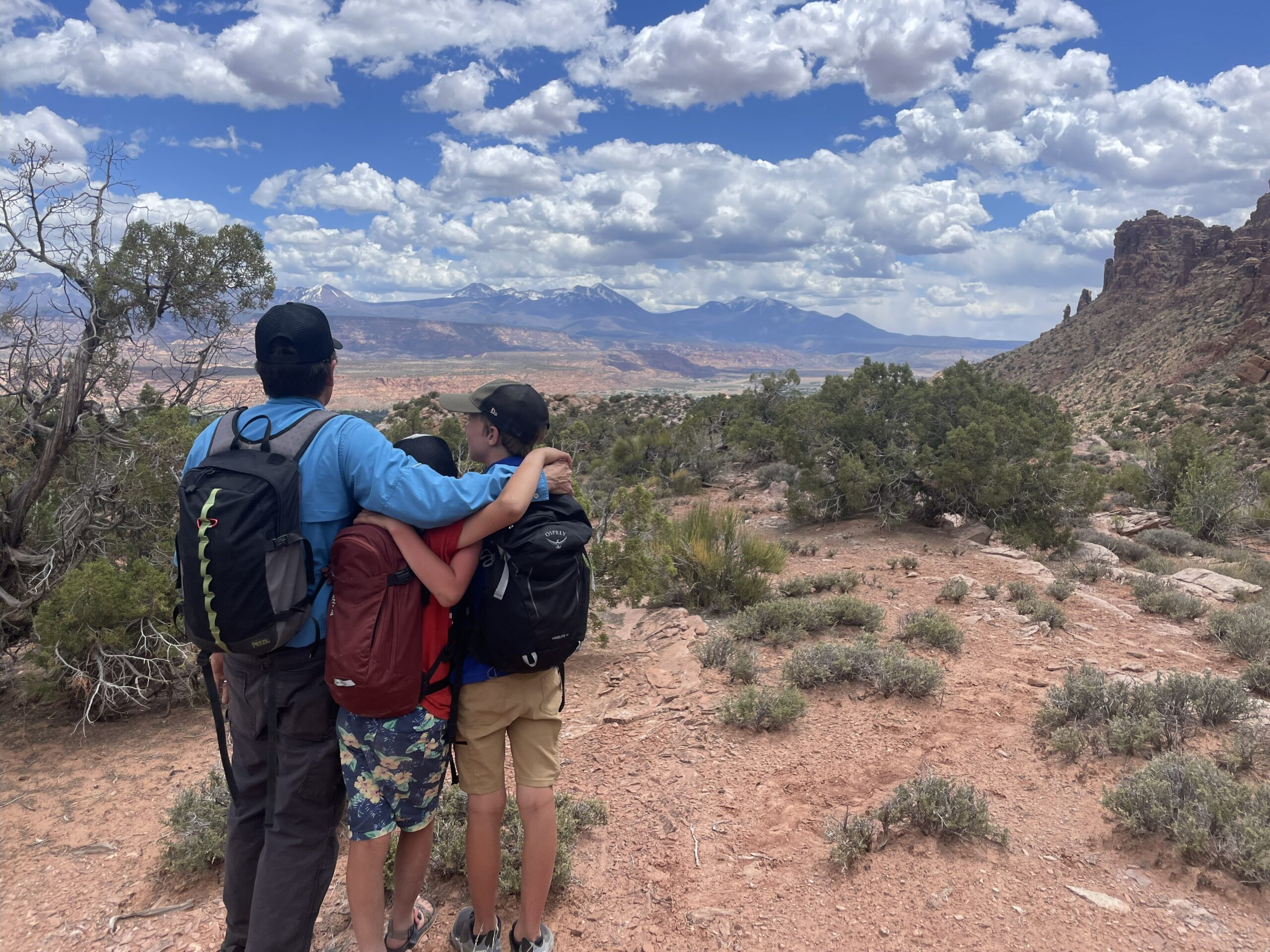 A father and two children stand arm-in-arm on a rocky desert overlook, gazing toward distant mountains under a partly cloudy sky. They wear backpacks and outdoor gear, symbolizing adventure, connection, and shared exploration in a rugged canyon landscape.