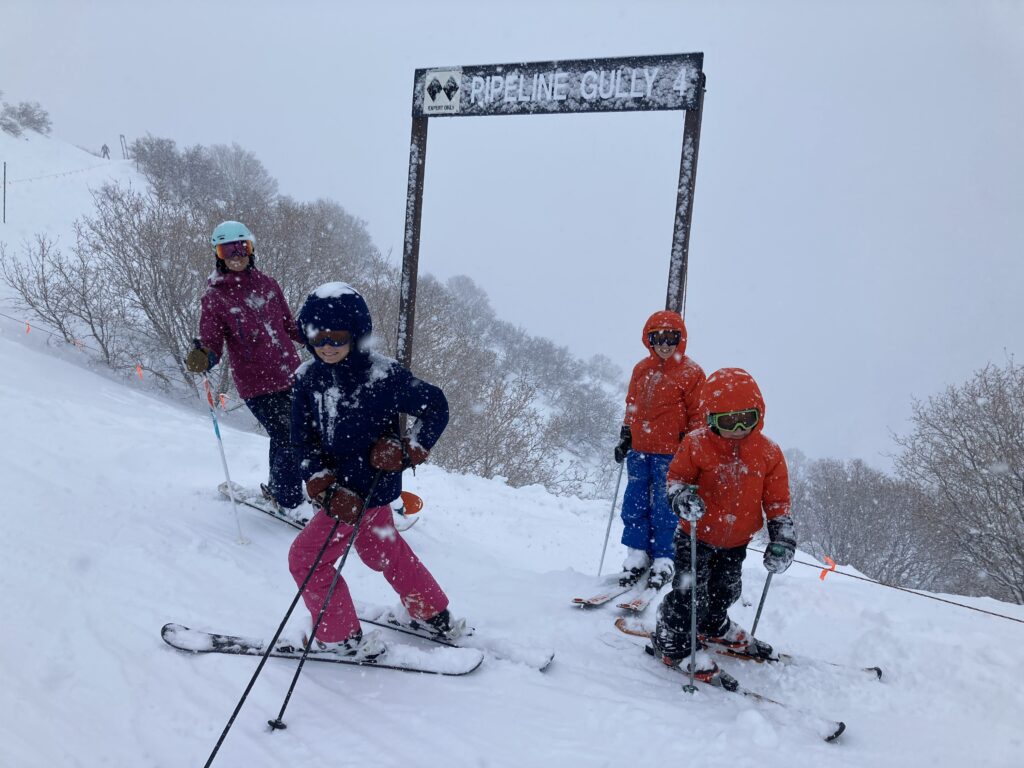 Four skiers pause near a snowy trail sign labeled 'Pipeline Gully 4' at Sundance Mountain Resort, sharing a moment of presence and togetherness in the falling snow — a snapshot of intentional family time on the mountain.