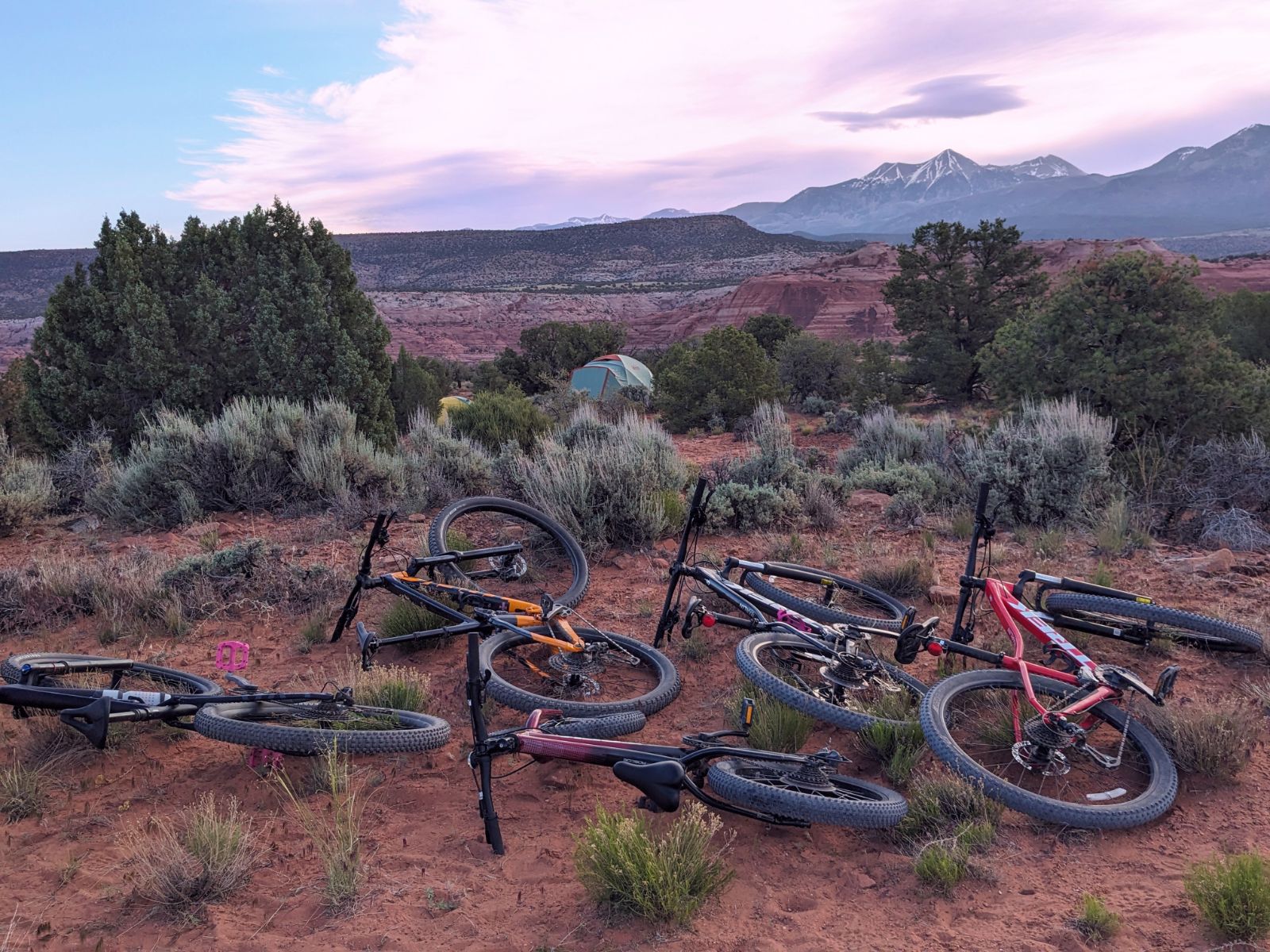 Mountain bikes rest on reddish desert soil near a tent nestled among trees, with red rock formations and snow-capped mountains rising in the distance under a softly lit sky. The scene evokes outdoor adventure, camping, and the quiet beauty of nature at sunrise or sunset.