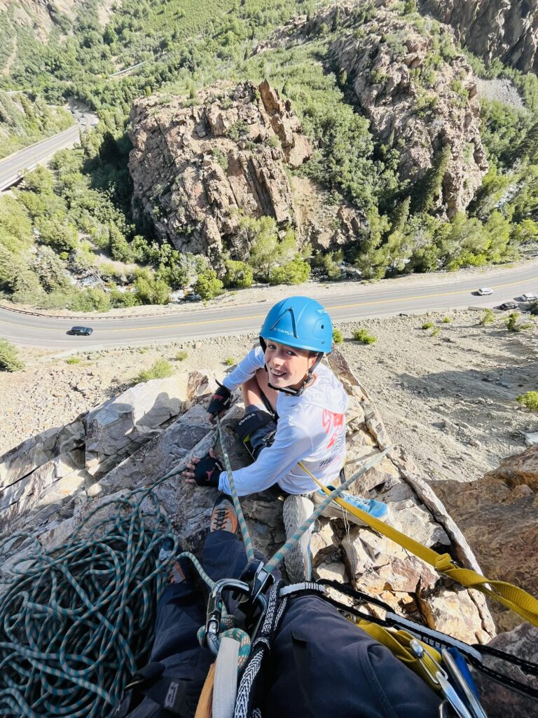 A child taking a quiet pause on a narrow ledge, safely tied in, experiencing the kind of challenge that forges connection.