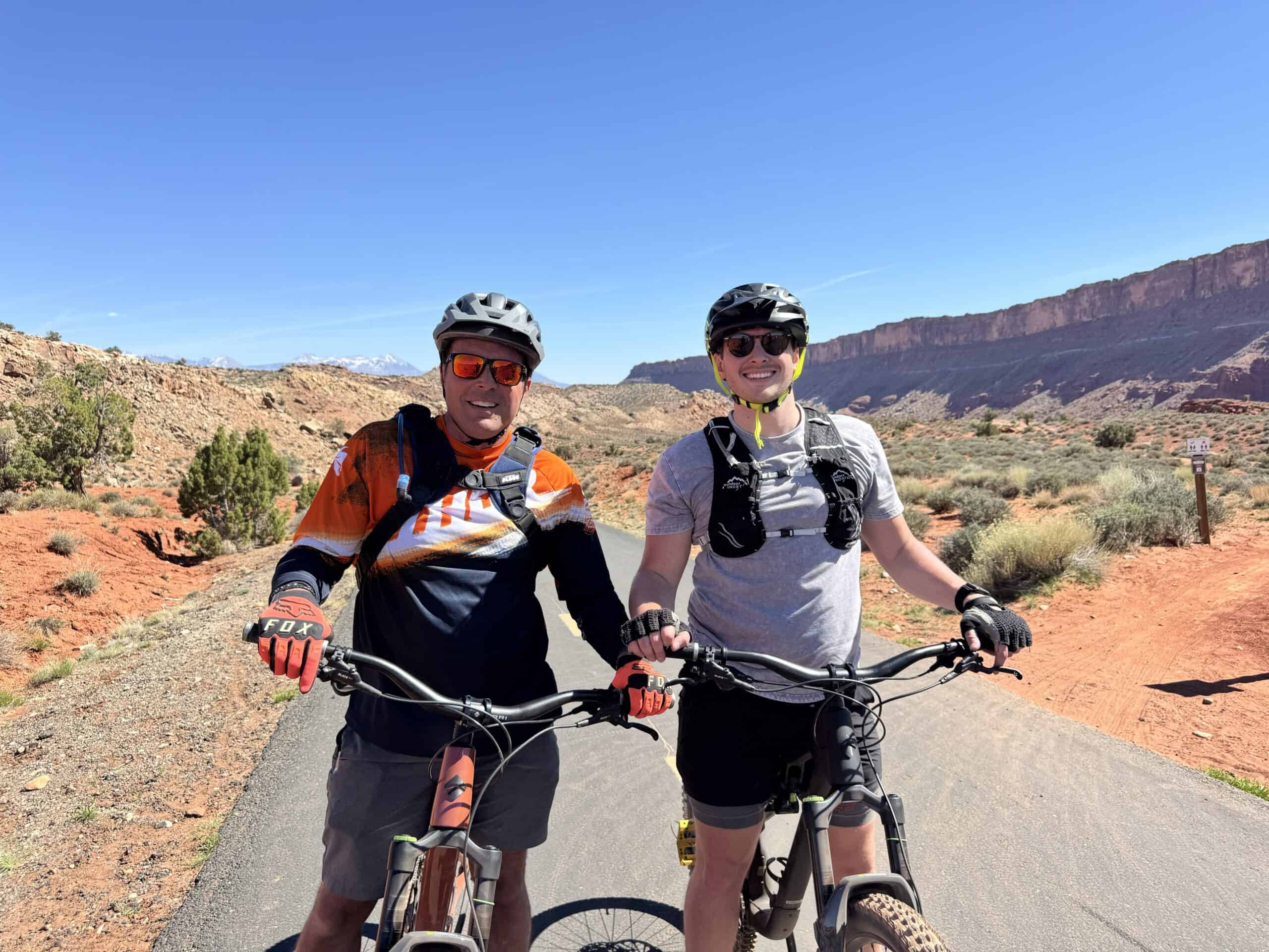 Two mountain bikers — an older and a younger rider — pause side by side on a paved trail, helmets on and bikes between them, with red sandstone cliffs and the La Sal Mountains in the background.