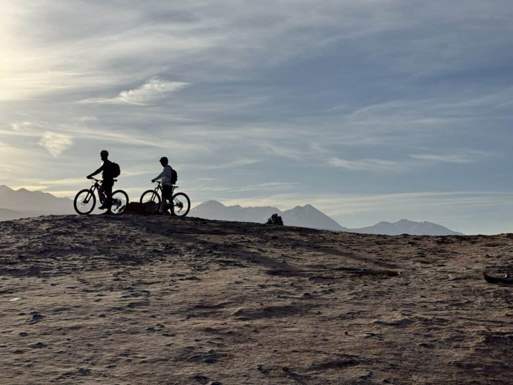 Two mountain bikers pause side by side on a slickrock ridge in Moab, silhouetted against a soft sky with the La Sal Mountains stretching across the horizon.