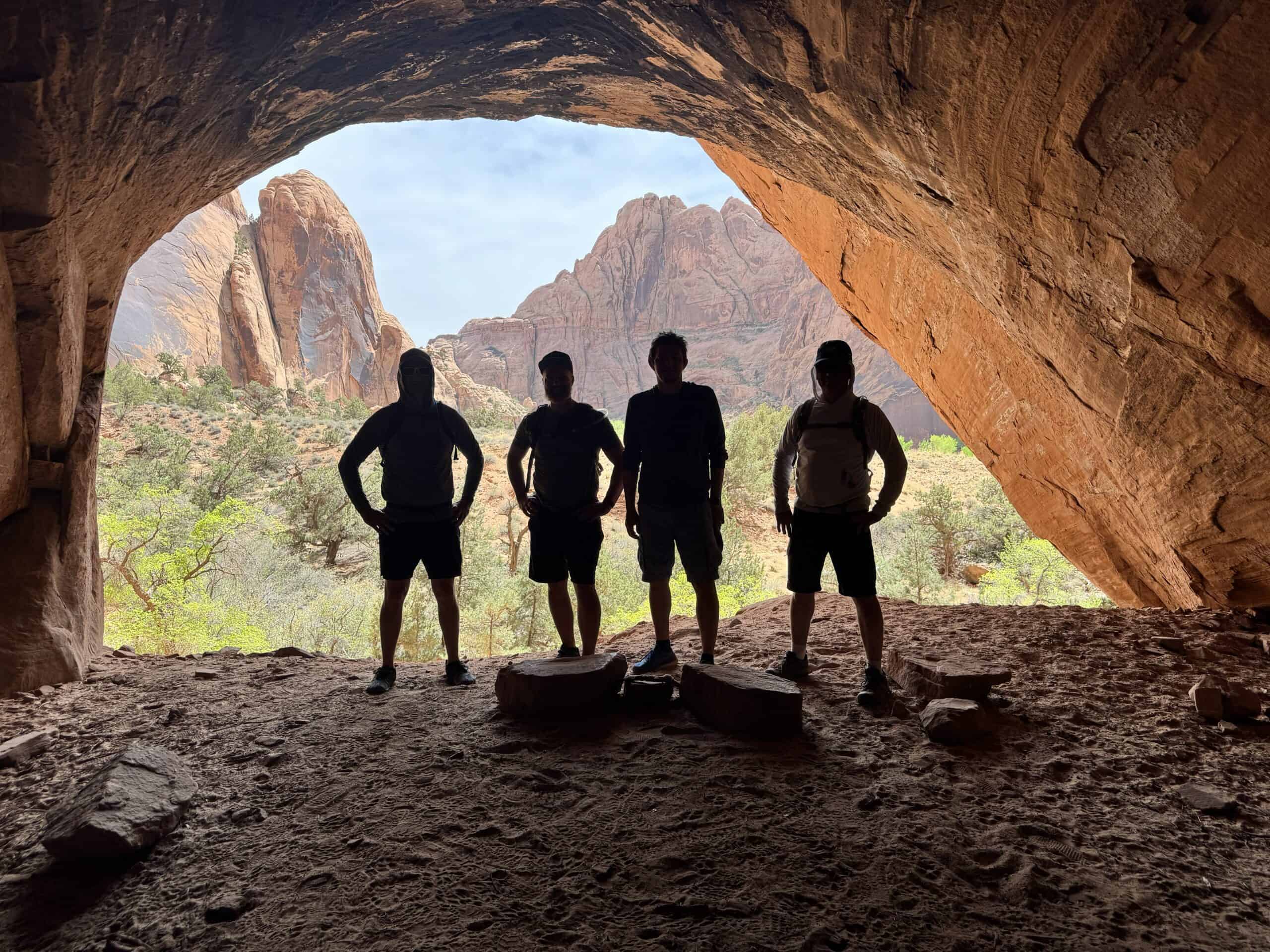 Four men stand together at the entrance of a sandstone alcove in Moab, silhouetted against the bright canyon beyond. Towering red rock formations and desert vegetation fill the view through the cave opening.