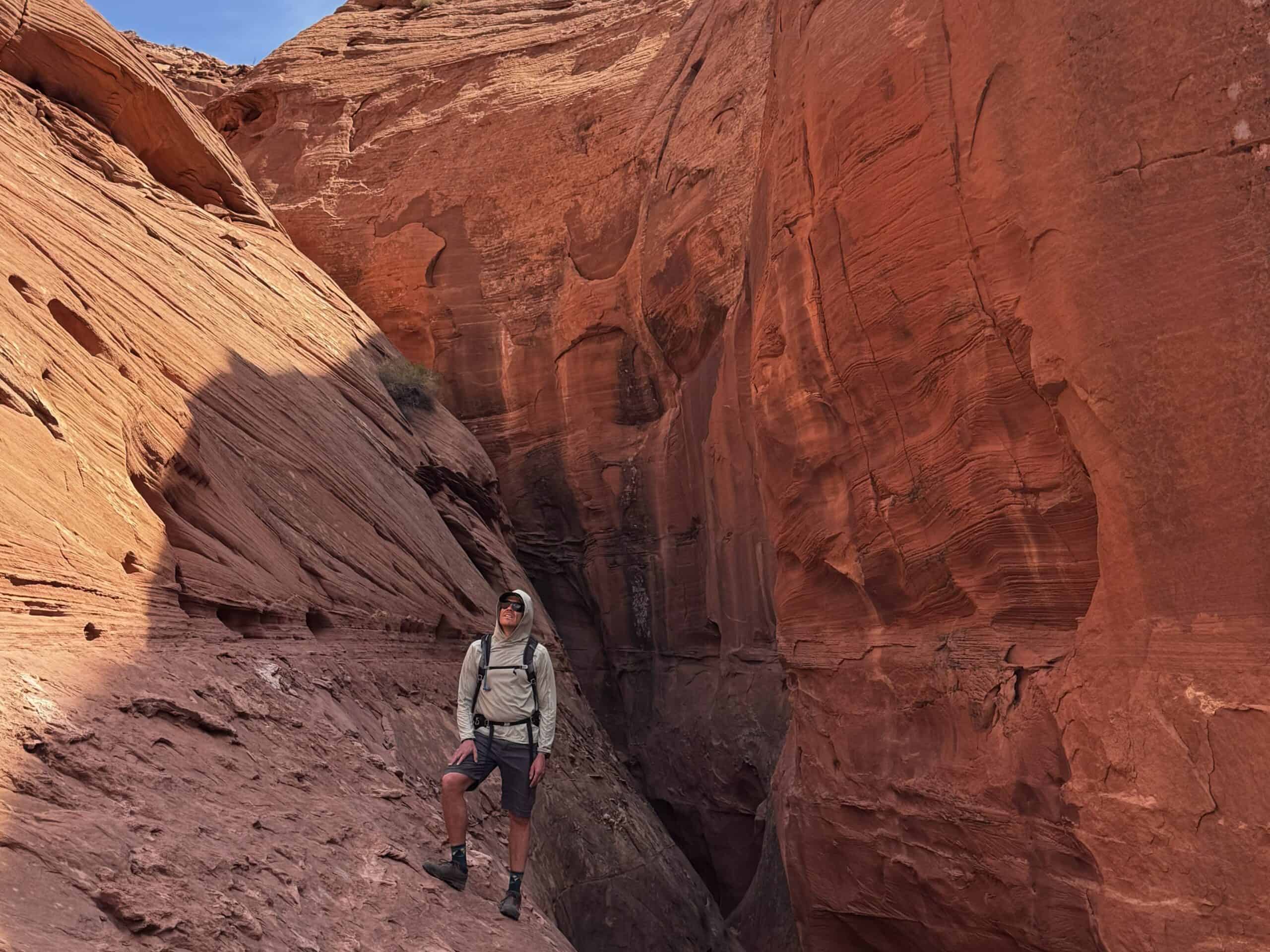A hiker pauses at the mouth of a slot canyon, looking up at the towering red sandstone walls that rise on either side and narrow into shadow behind him.