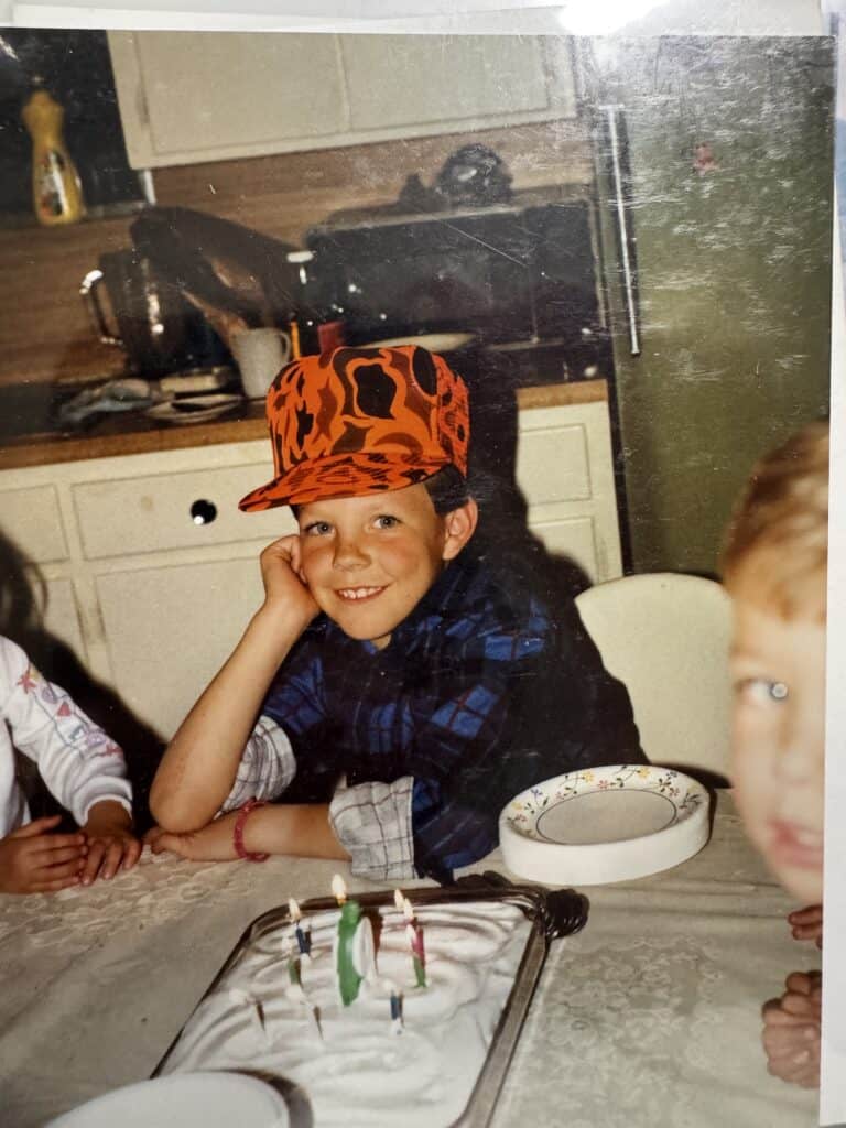 The author as a boy in his grandmother's kitchen celebrating his birthday, wearing an orange camo cap and blue plaid shirt behind a candlelit cake.