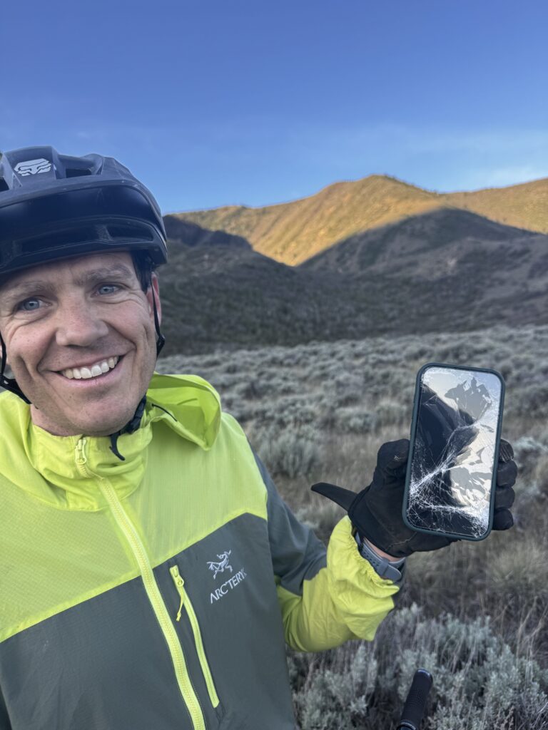 Smiling mountain biker in helmet and neon jacket holds up a phone with a shattered screen on a sagebrush trail at sunrise.